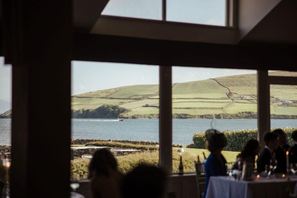 View through large windows showing people seated at tables inside, with a scenic backdrop of a lake and green hills under a clear sky outside.