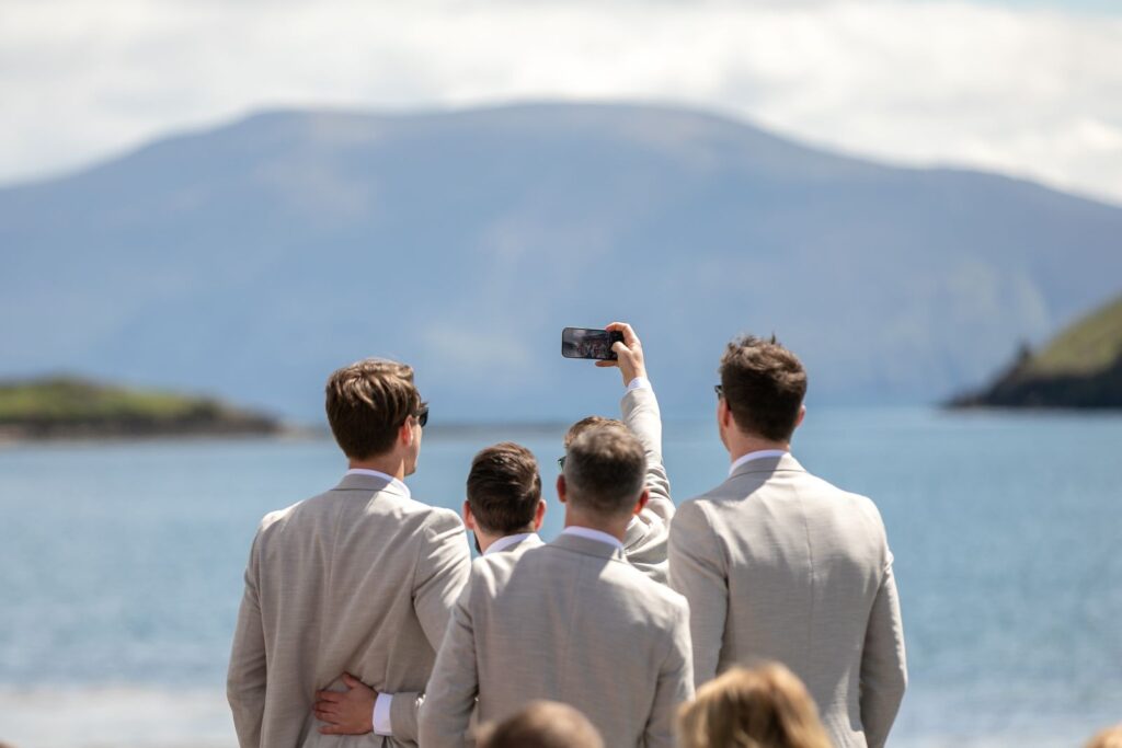 Four men in light suits stand by the water, with one holding up a phone to take a selfie; mountains and sky are visible in the background.