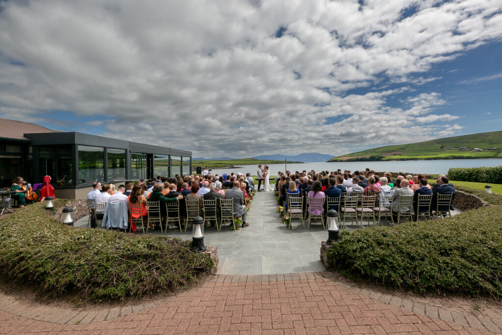A large group of people sits outdoors facing a couple at an altar during a wedding ceremony, with green hills and a cloudy sky in the background.