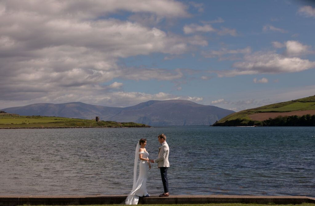 A bride and groom stand facing each other by a large body of water, with green hills and mountains in the background under a partly cloudy sky.