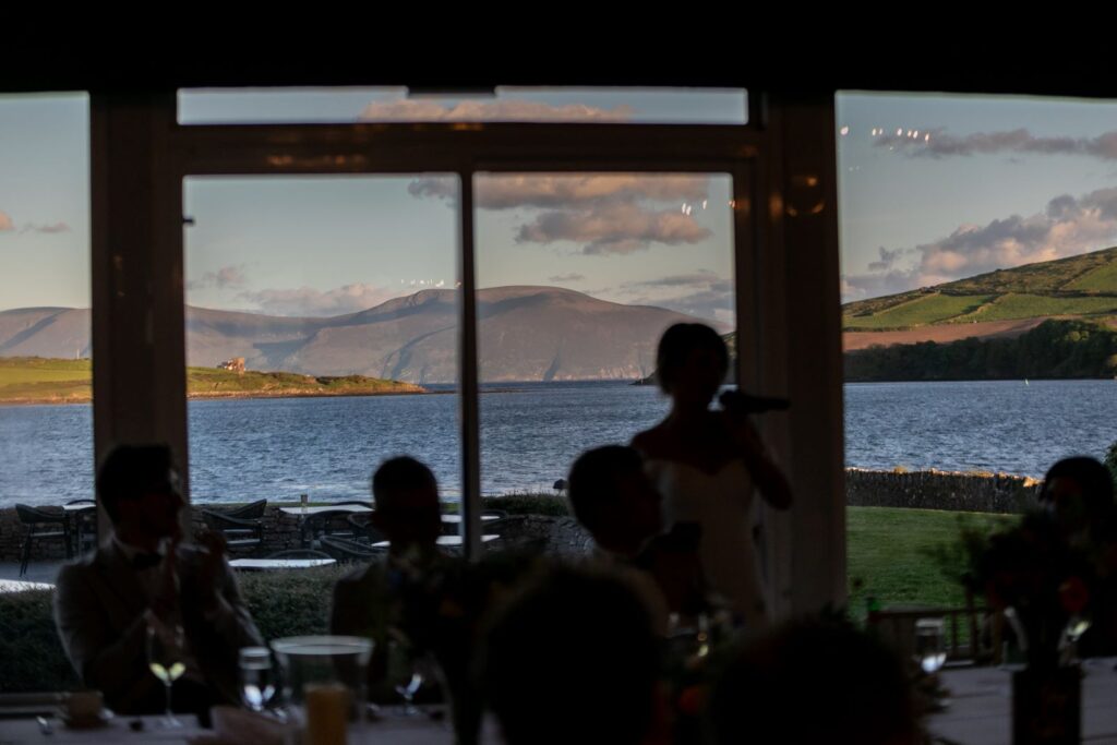 A group of people sit indoors at a table near a large window overlooking a lake and distant hills, with a person standing and speaking into a microphone.