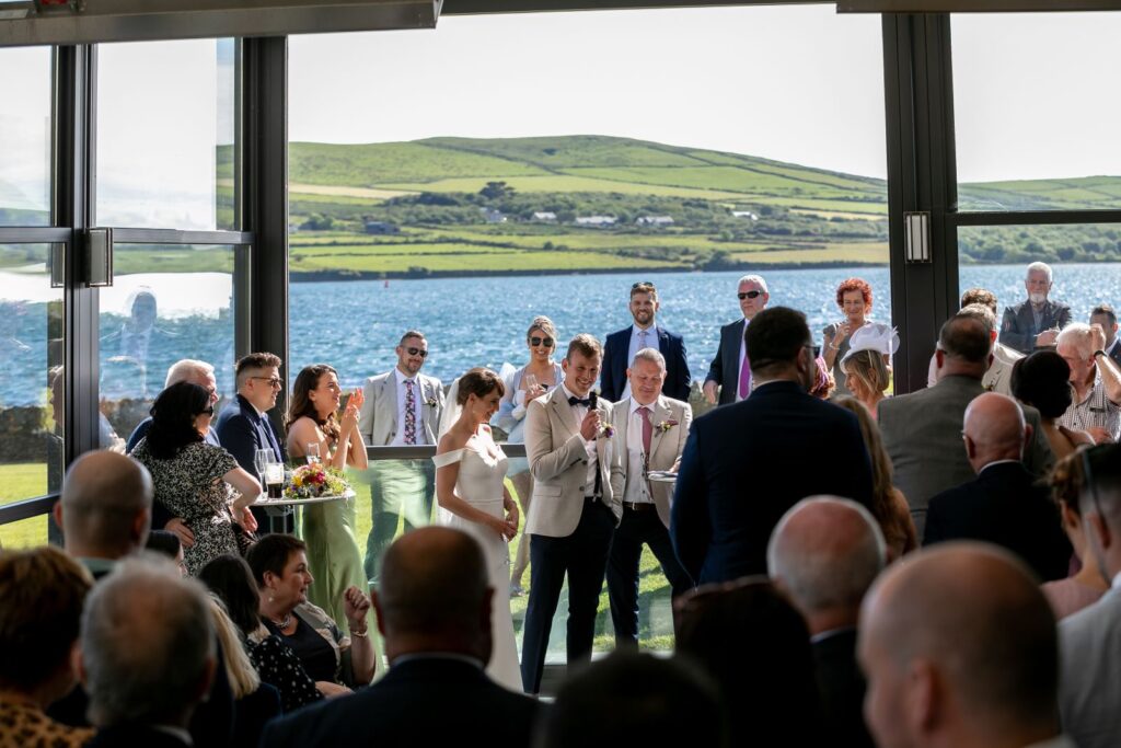 A bride and groom stand together holding a microphone, addressing wedding guests indoors with large windows showing a scenic waterfront and rolling green hills outside.
