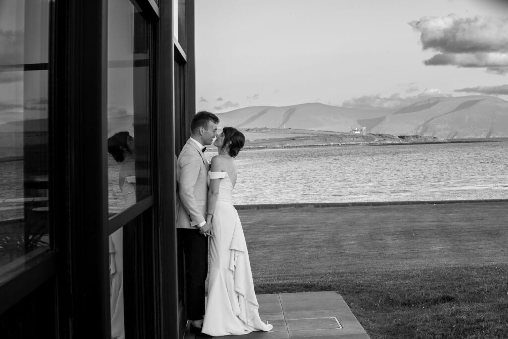 A bride and groom stand close together outside a building, overlooking water and distant mountains on a grassy landscape.