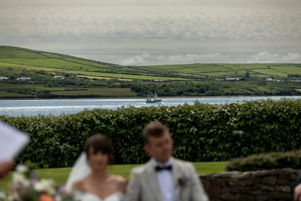 A boat sails on a river with green hills and houses in the background; a blurred bride and groom are in the foreground.