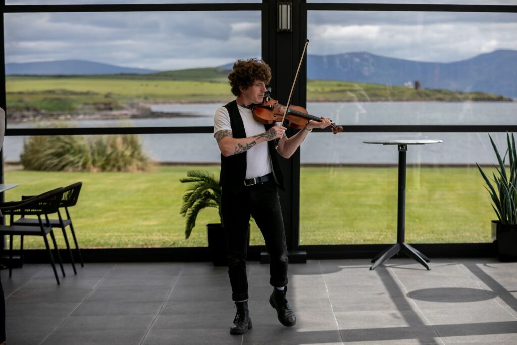 A person with curly hair plays a violin indoors, standing by large windows overlooking a grassy landscape and water with hills in the background.