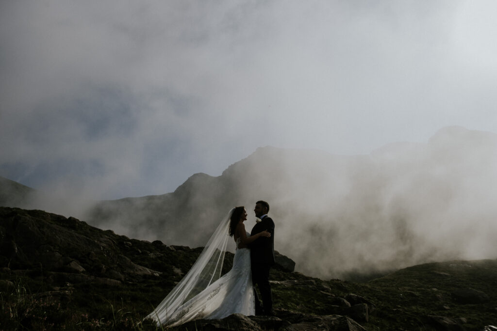 A bride and groom stand together on rocky terrain with mist and mountains in the background.