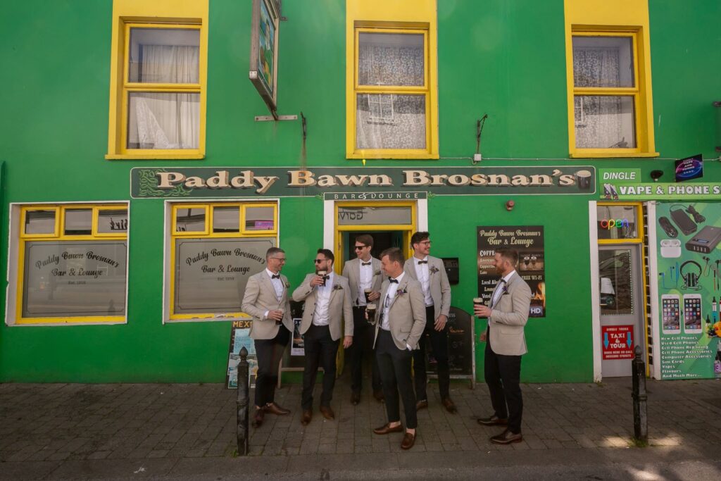 Five men in suits stand and talk outside Paddy Bawn Brosnan's Bar & Lounge, a green building with yellow trim, on a sunny day.