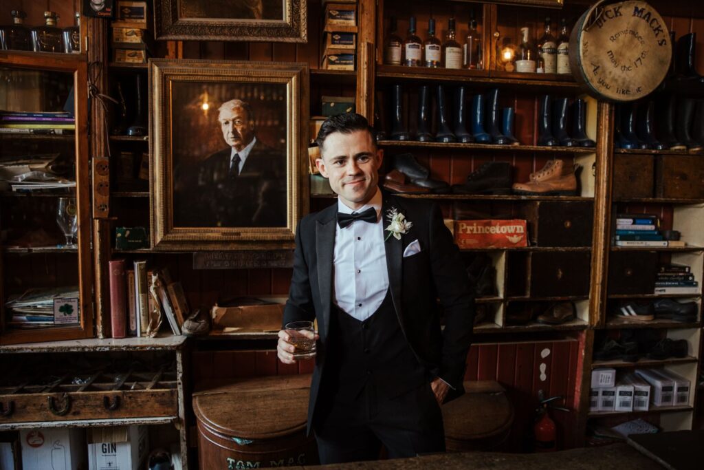 A man in a tuxedo holds a drink and stands in a vintage room with shelves of boots, bottles, books, and a large framed portrait behind him.
