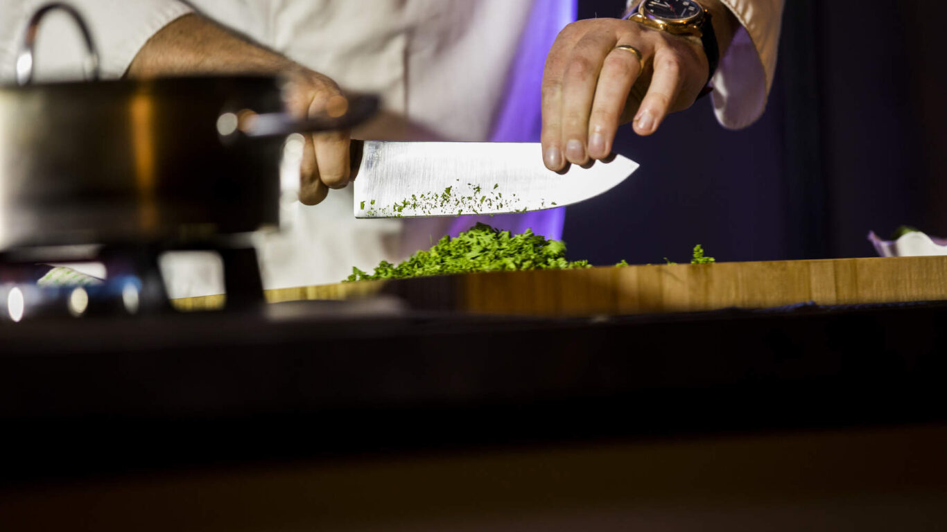 A person in a chef's coat chops fresh herbs on a wooden board with a large knife; a pot is visible on the left side of the image.