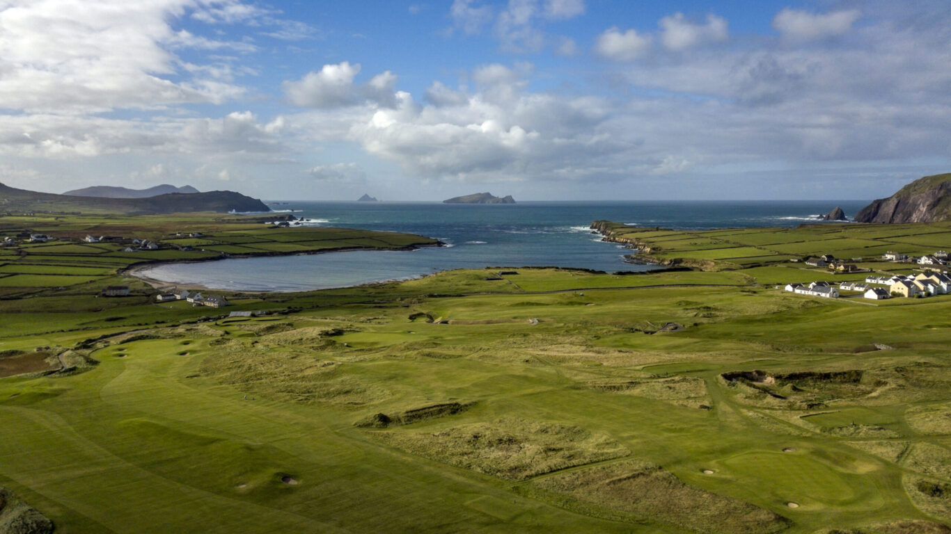 Aerial view of a coastal landscape with green fields, a bay, scattered houses, and distant islands under a partly cloudy sky.