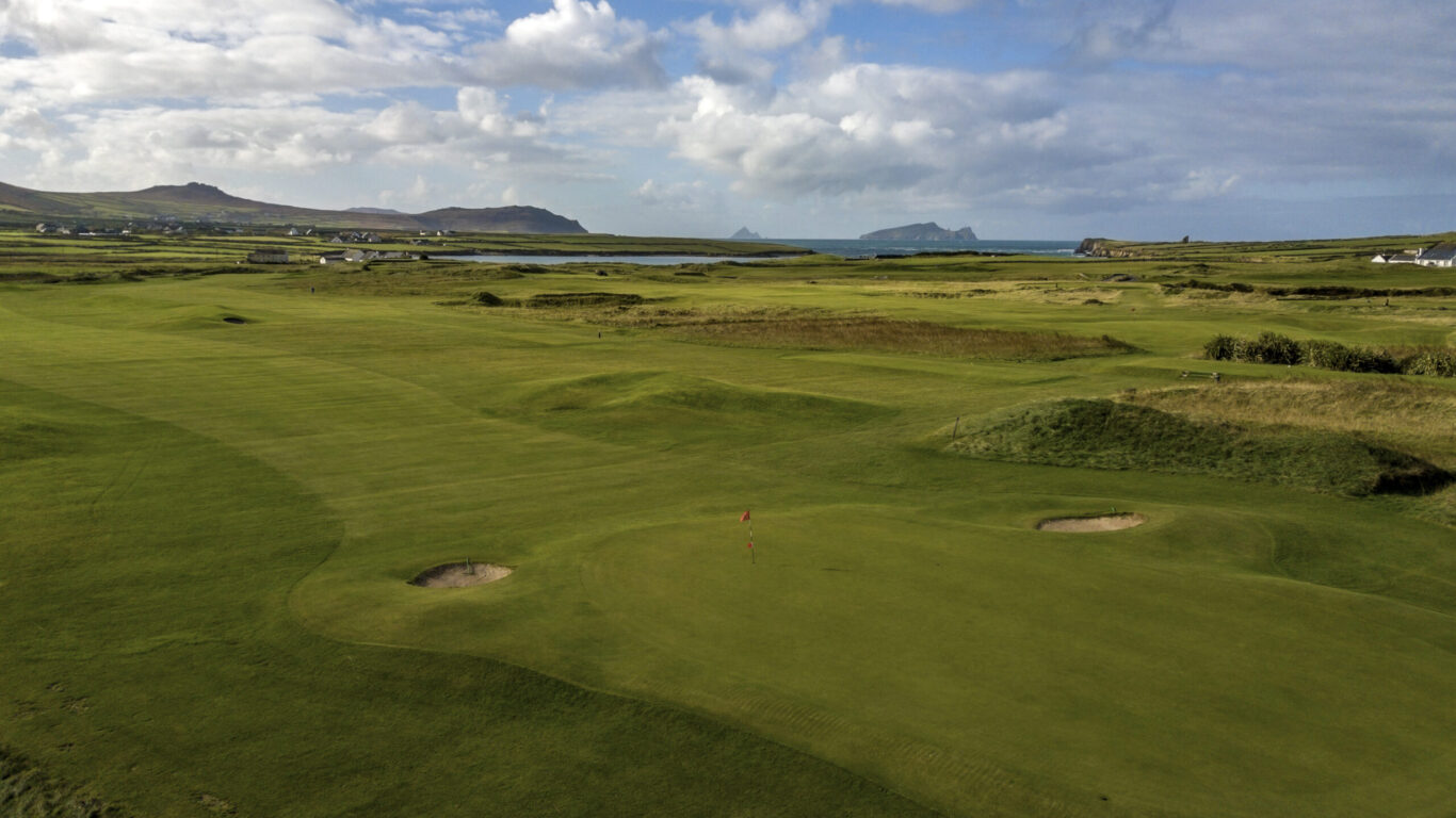 Aerial view of a green golf course with sand bunkers, surrounded by open landscape, distant hills, and a partly cloudy sky.