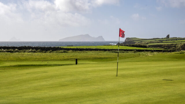 A red golf flag stands on a green golf course overlooking the ocean, with distant islands and a partly cloudy sky in the background.