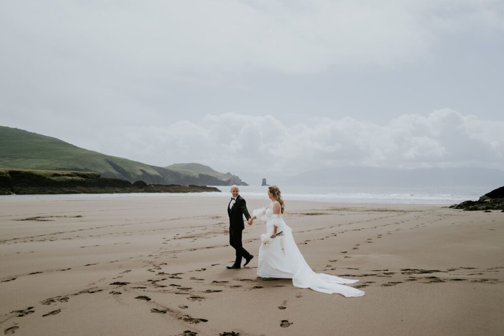 A bride and groom walk hand in hand across a sandy beach, with footprints behind them and distant hills under a cloudy sky.