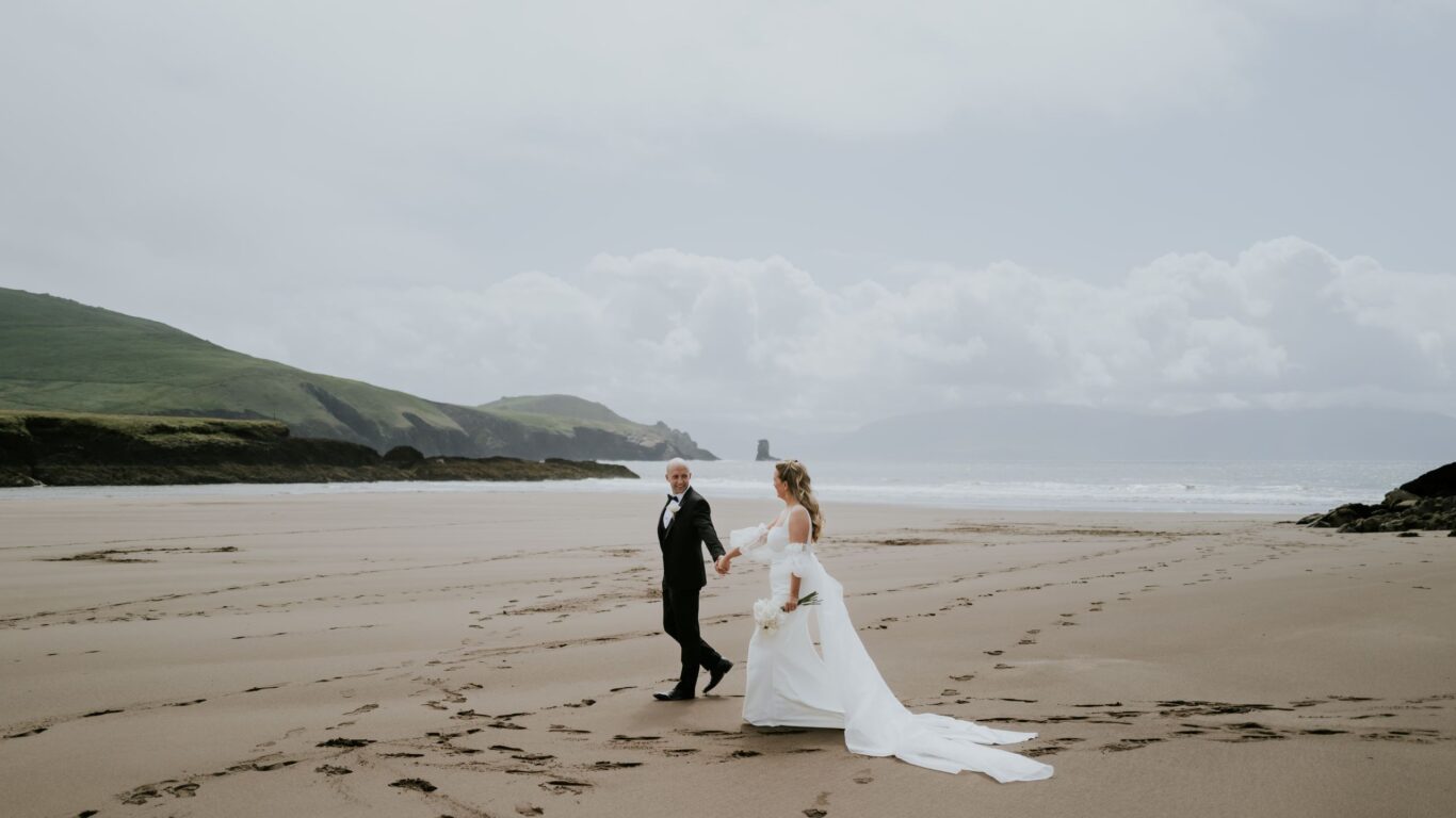 A bride and groom walk hand in hand across a sandy beach, with footprints behind them and distant hills under a cloudy sky.