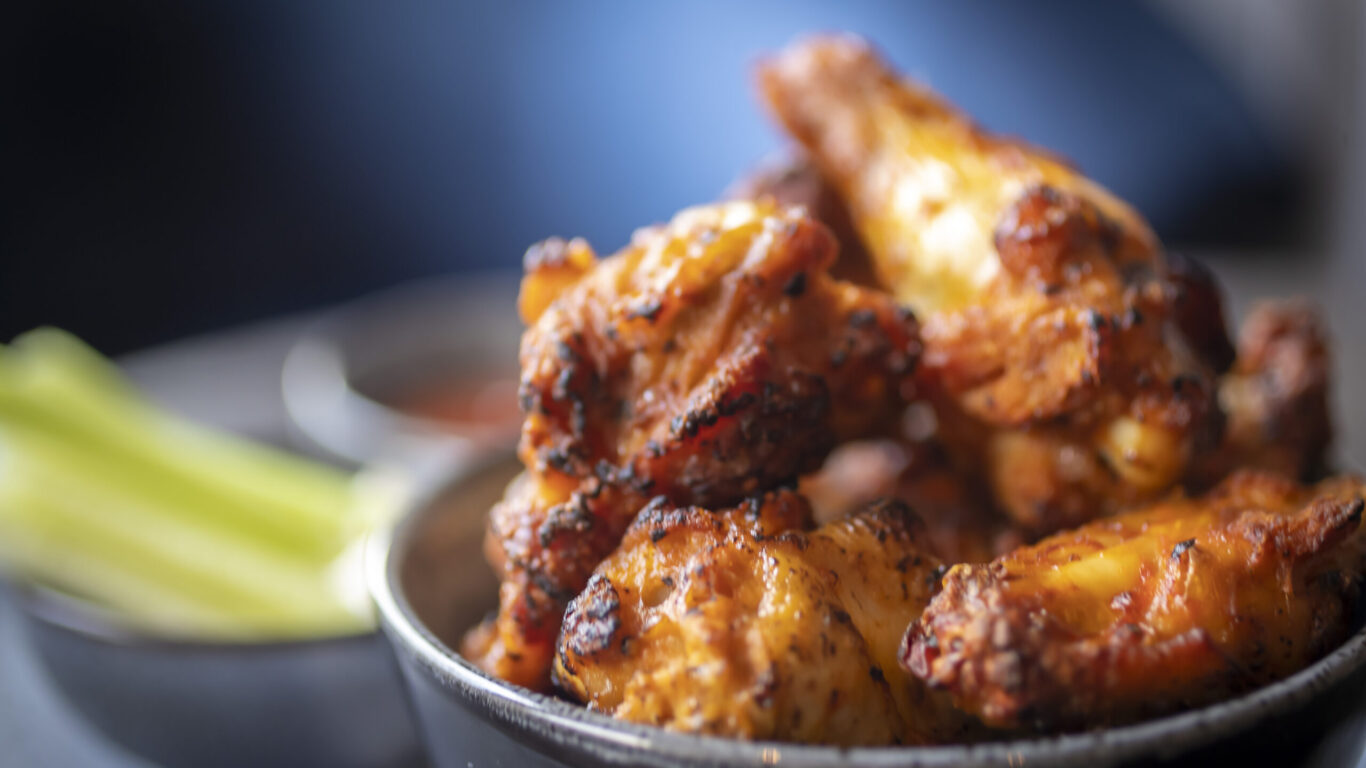 A close-up of a bowl filled with grilled chicken wings, with a side of celery sticks and dipping sauce in the background.