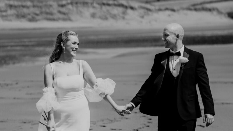 A bride and groom walk hand in hand on a sandy beach, dressed in formal wedding attire, with mountains in the background.