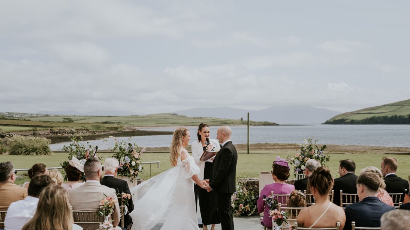 A couple stands with an officiant at an outdoor wedding ceremony by a lake, with seated guests on either side of a red carpet under a partly cloudy sky.