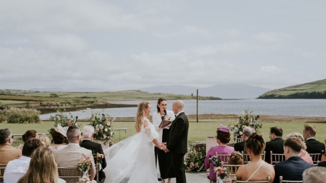 A couple stands with an officiant at an outdoor wedding ceremony by a lake, with seated guests on either side of a red carpet under a partly cloudy sky.