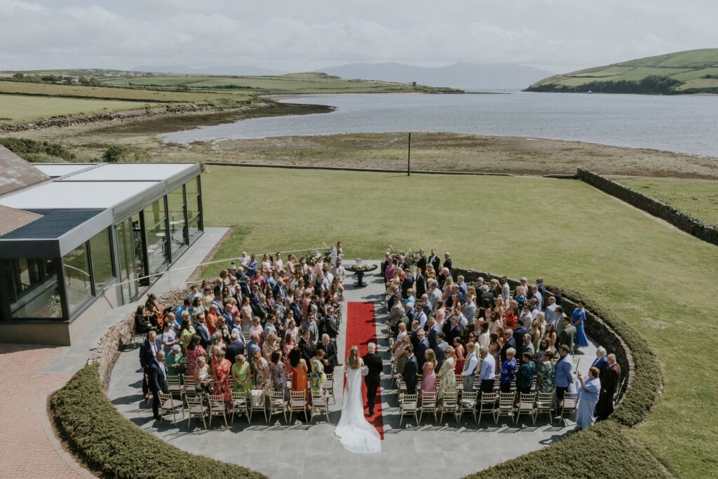 A wedding ceremony taking place outdoors by the water, with guests seated in rows around a red carpet leading to the couple standing at the altar.