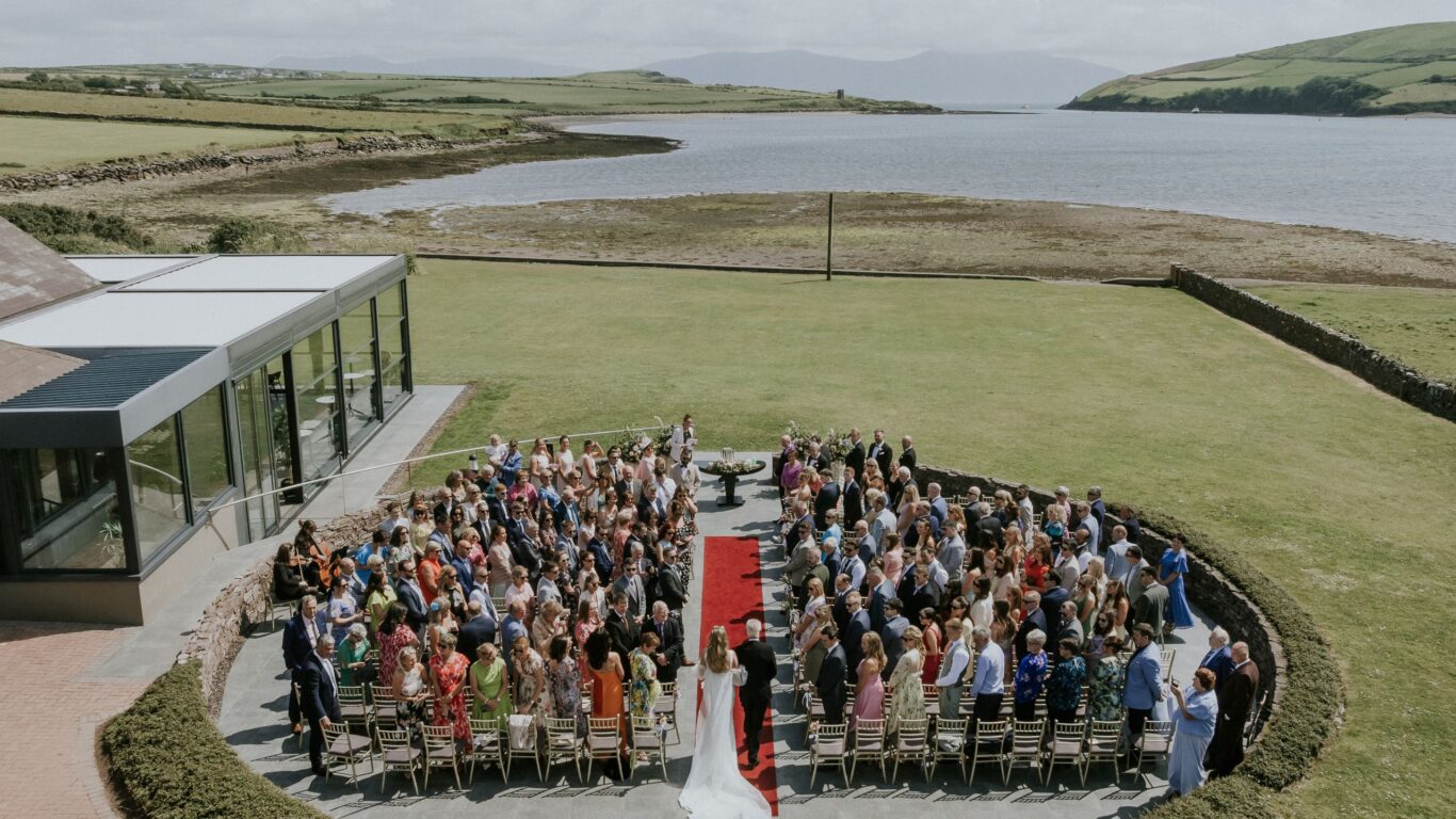 A wedding ceremony taking place outdoors by the water, with guests seated in rows around a red carpet leading to the couple standing at the altar.
