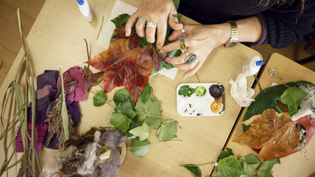 A person arranges autumn leaves and craft materials on a table, with paint, glue, and various art supplies visible.