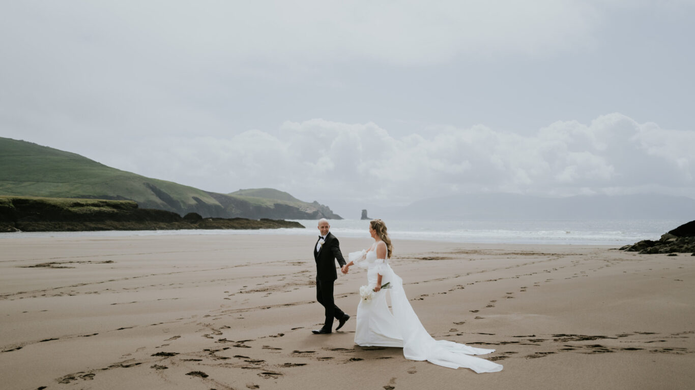 A bride and groom walk hand in hand across an empty beach, with hills and cloudy skies in the background.