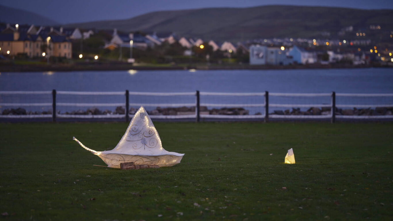 Two illuminated paper sculptures resembling boats sit on a grassy area by a waterfront, with houses and hills visible in the background at dusk.