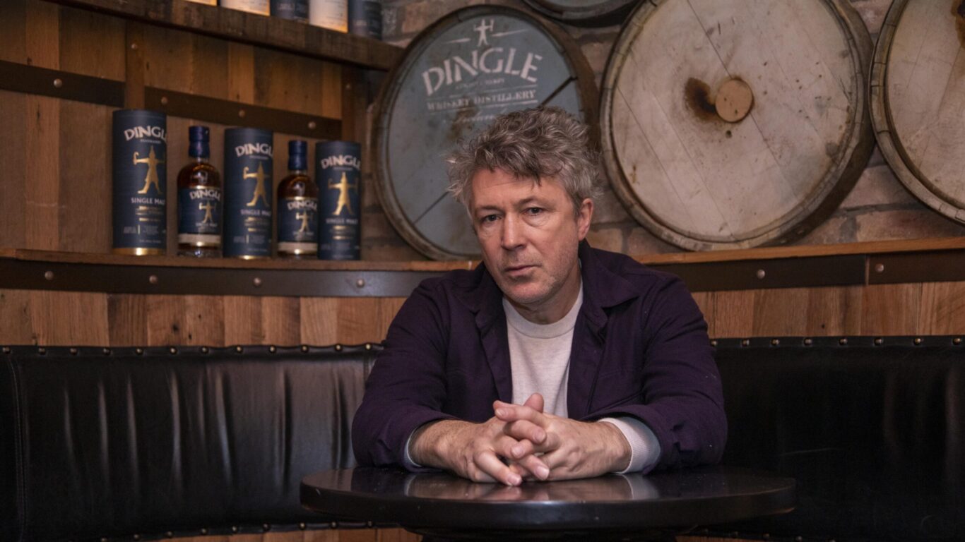 A man sits at a round table in a bar with wooden barrels and bottles of Dingle whiskey displayed on shelves behind him.