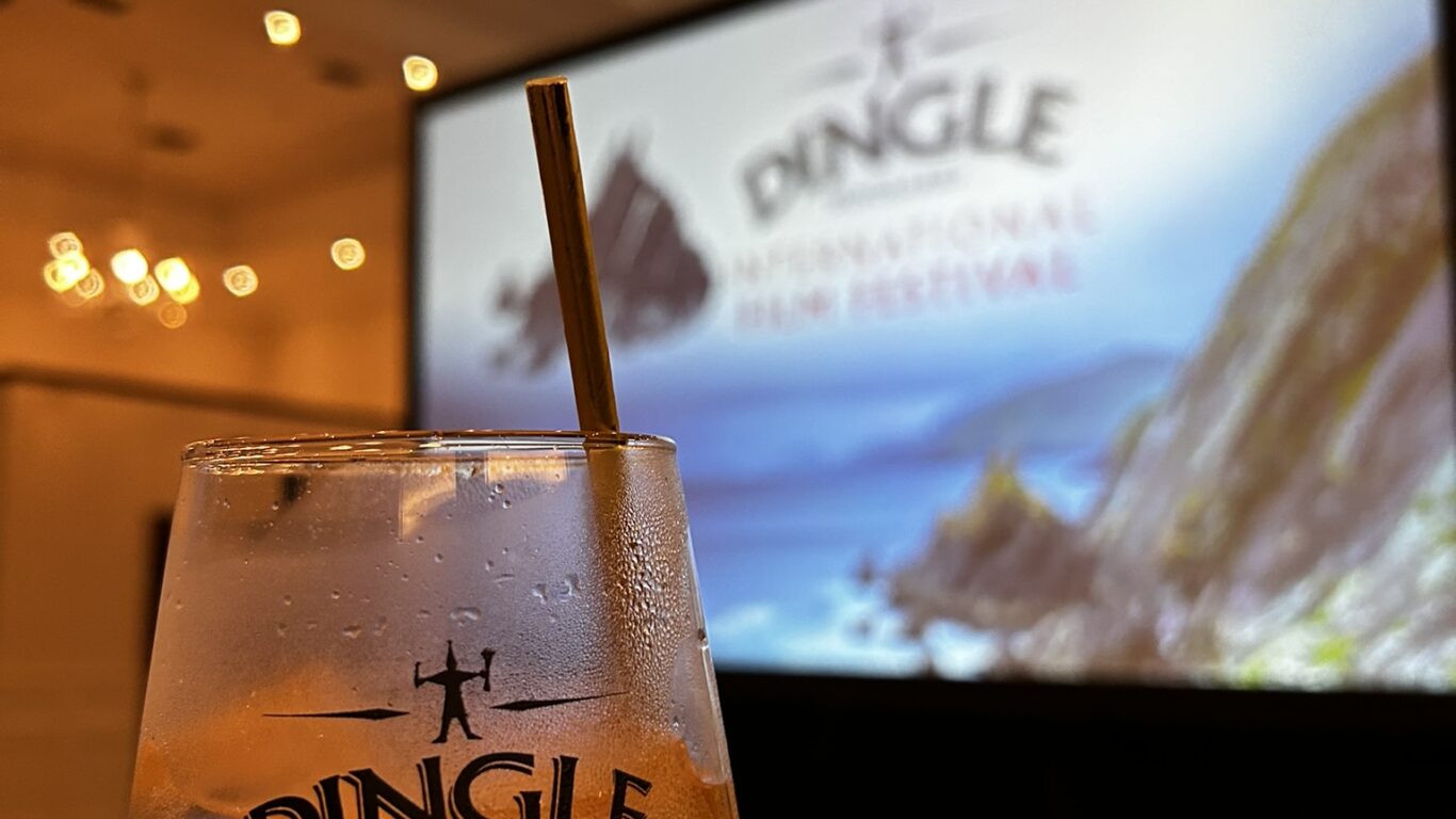 A hand holds a Dingle Distillery glass with a drink in front of a large screen displaying the Dingle International Film Festival logo in a well-lit event space.