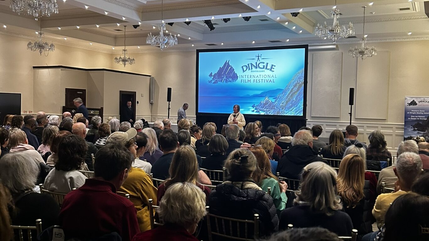 A large audience sits facing a stage with a screen displaying