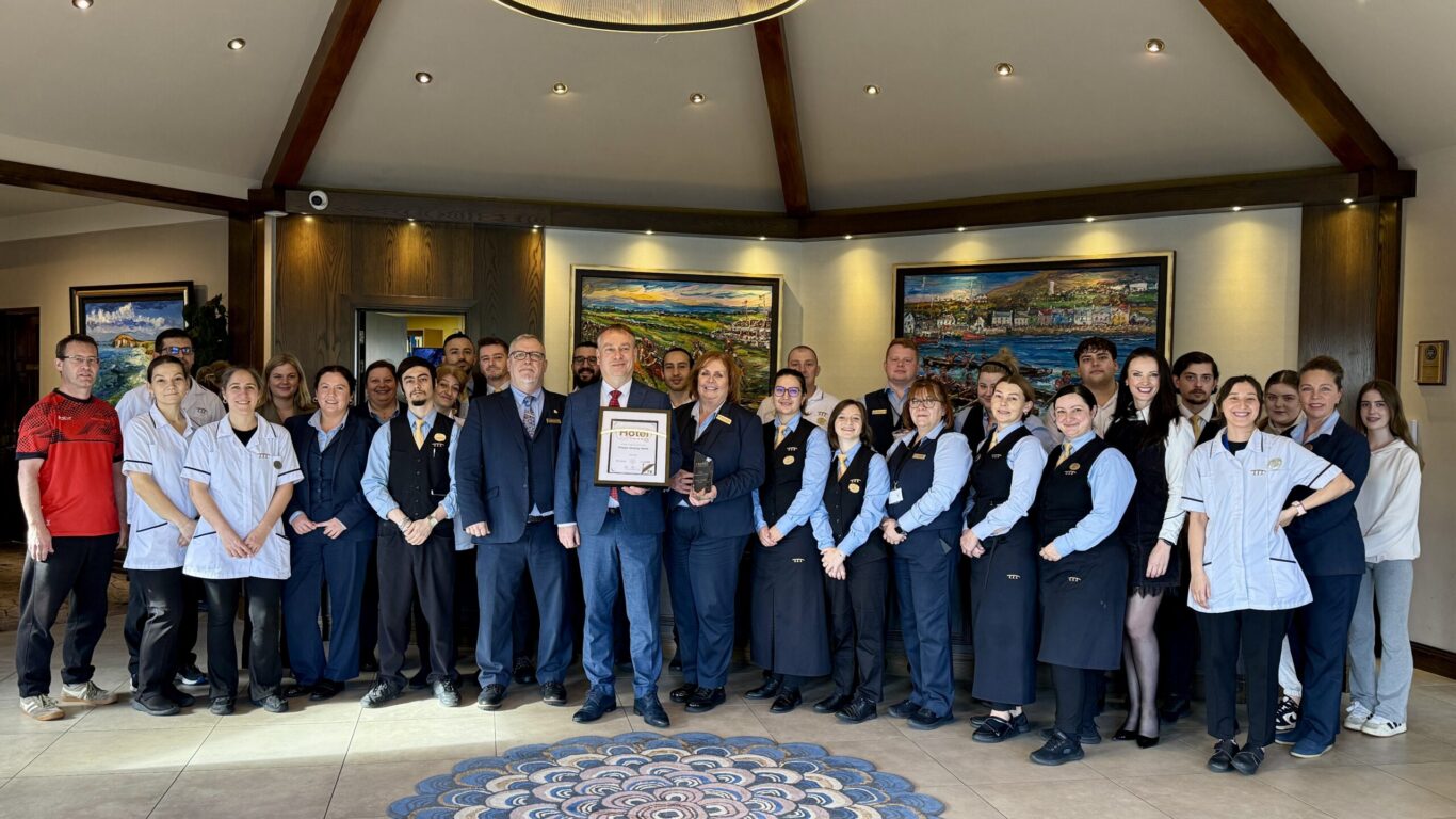 A large group of professionally dressed people stand together indoors, with one man in the center holding a framed certificate.