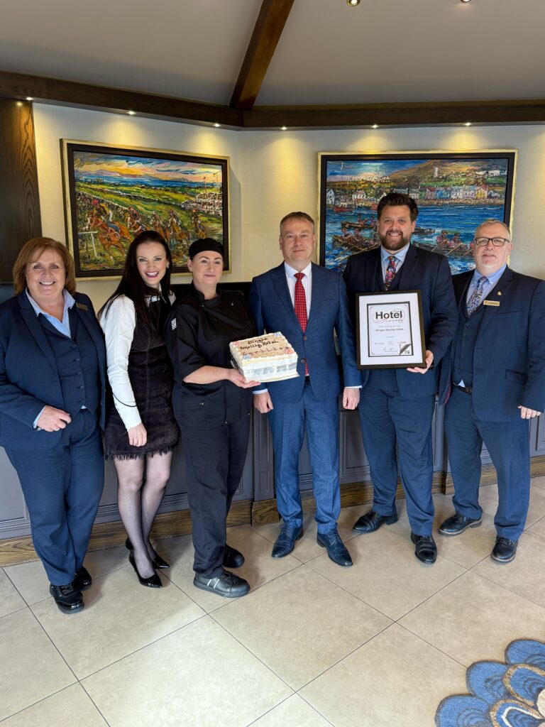 Six people in business attire stand indoors, with one person holding a certificate and another holding a box. Framed colorful paintings hang on the wall behind them.
