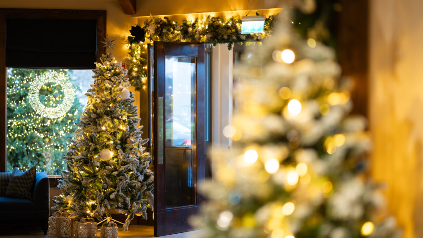 A decorated room with lit Christmas trees and garlands, presents under one tree, and a glass door showing a lit wreath outside.