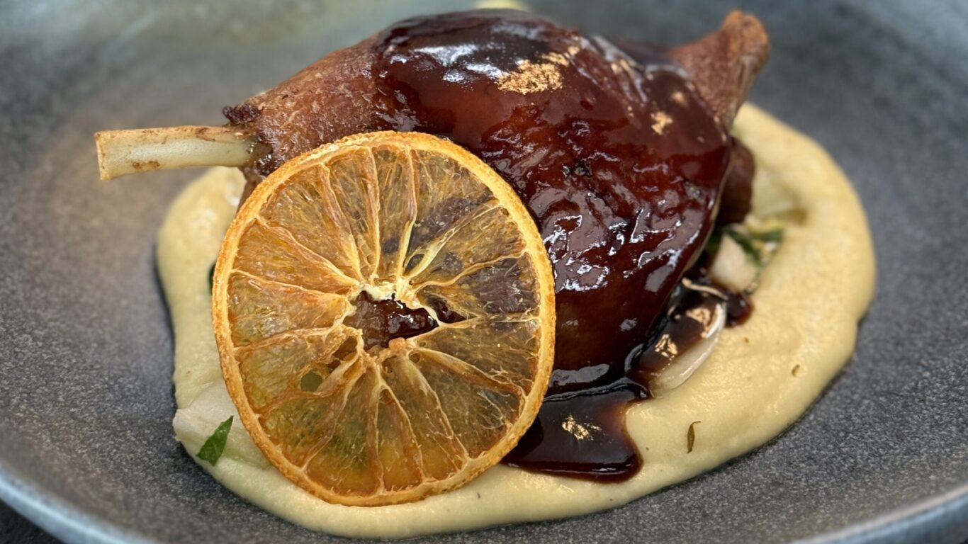 A plated dish of braised meat with a glossy dark sauce, served on creamy mashed potatoes and garnished with a slice of dried orange.