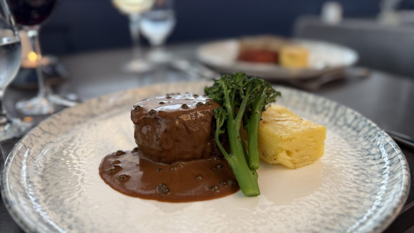 A plated meal featuring a steak with brown sauce, broccolini, and a rectangular portion of layered potatoes, with wine glasses in the background.
