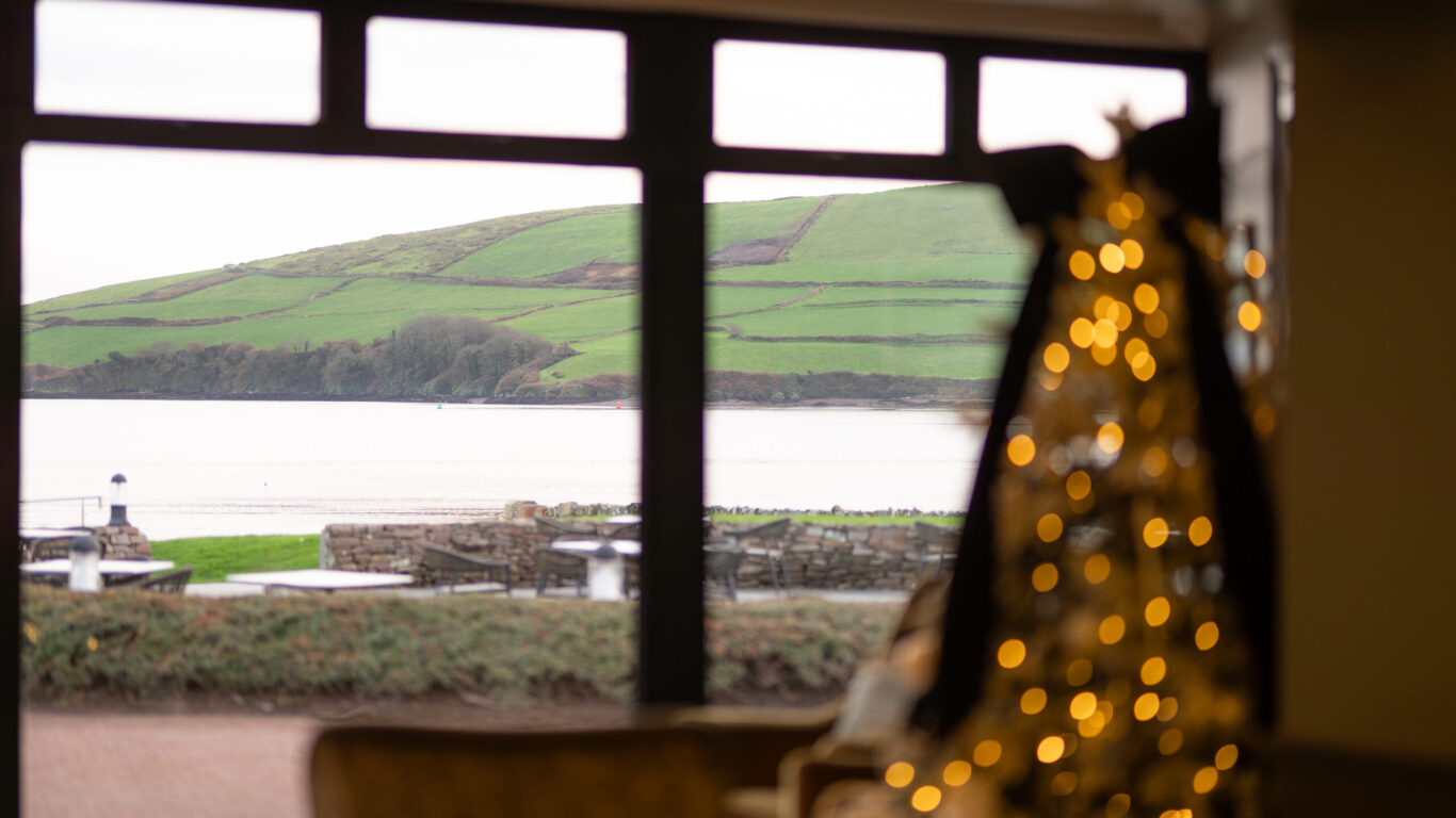 View of green hills and water seen through large windows, with an out-of-focus Christmas tree decorated with lights in the foreground.