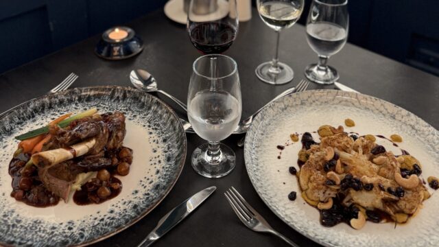 Two plates of food on a dark table: one with lamb shank and vegetables, the other with roasted cauliflower. Three wine glasses and a water glass are also on the table.