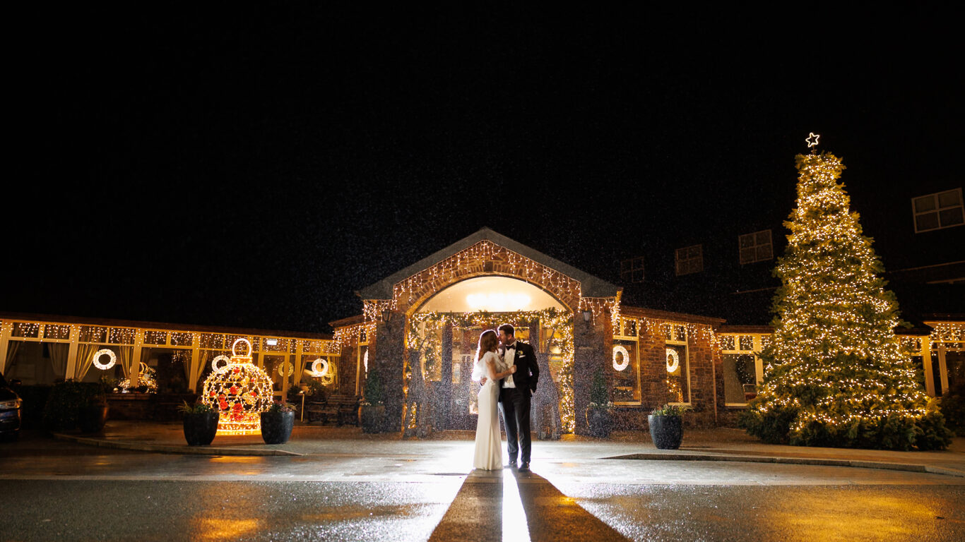 A bride and groom stand together at night in front of a building decorated with string lights and a large lit Christmas tree.