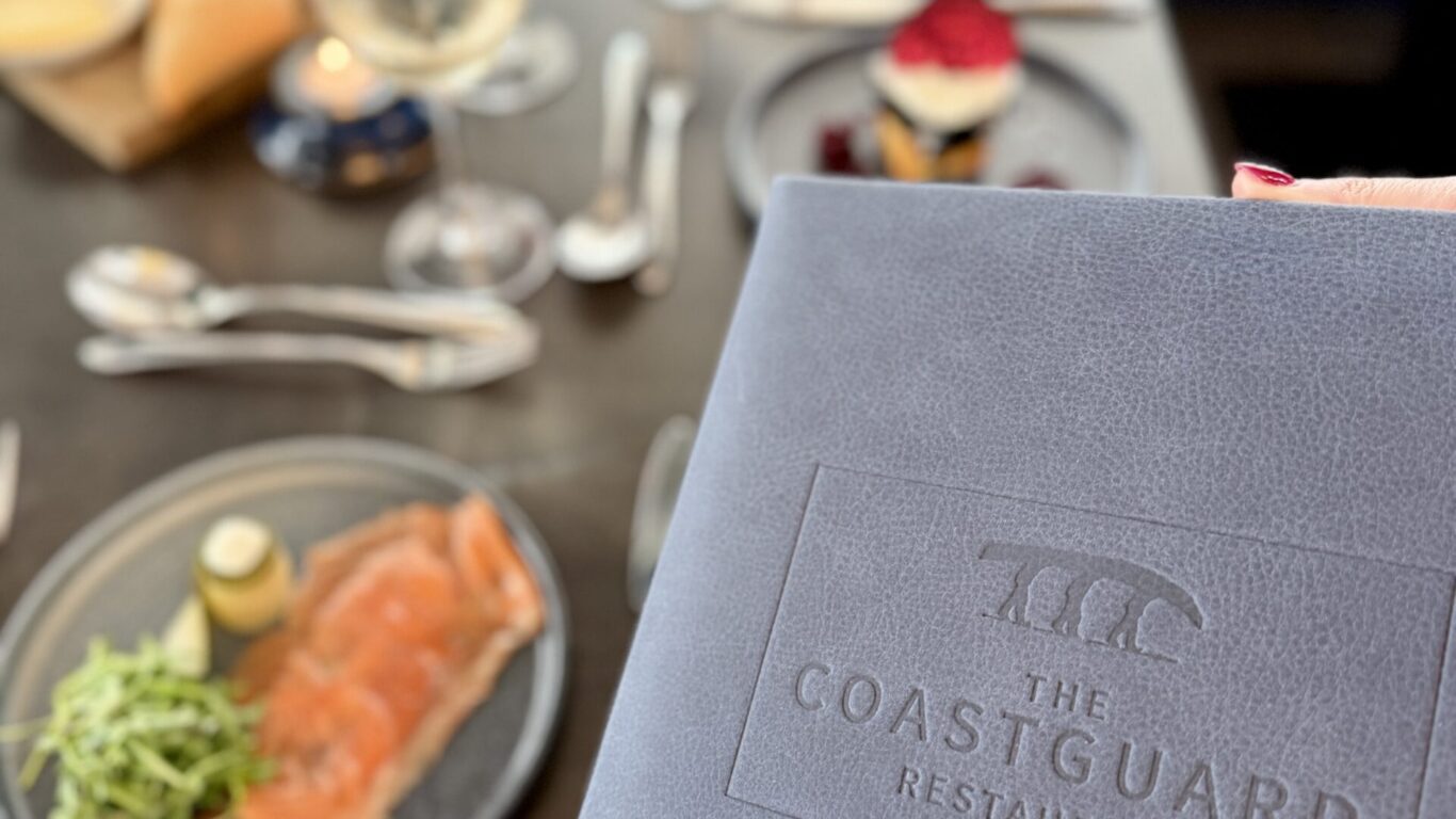 A close-up of a menu from The Coastguard Restaurant with a table set for a meal, including smoked salmon, dessert, and drinks in the background.
