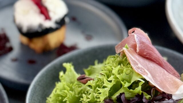 A bowl of mixed greens with figs, pecans, and prosciutto is in focus, with blurred plates of desserts and main courses in the background.