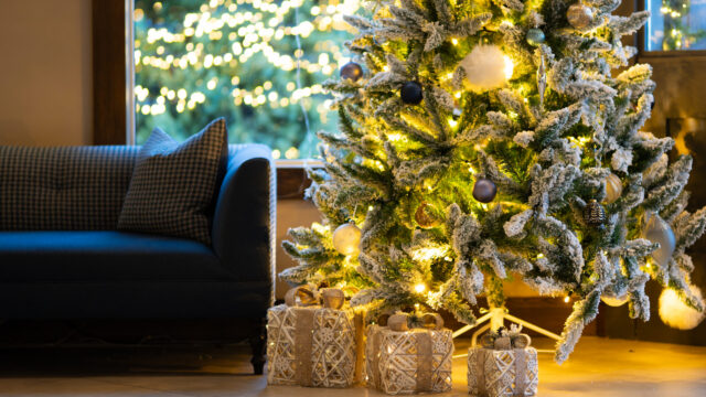 A decorated Christmas tree with lights and baubles stands by a window next to a blue sofa, with wrapped presents placed underneath.