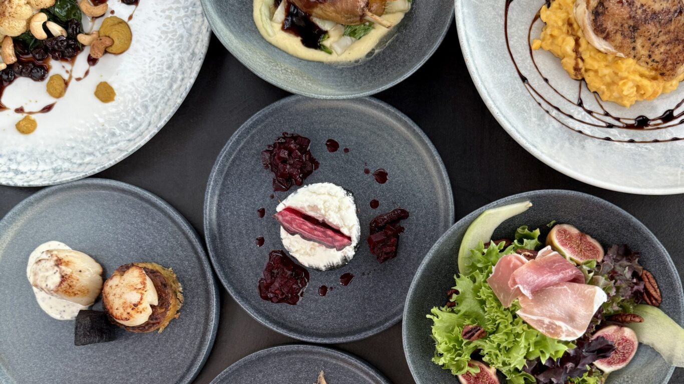 An overhead view of assorted gourmet dishes on gray plates, including salads, smoked salmon, meat entrees, and a dessert, arranged on a dark surface.