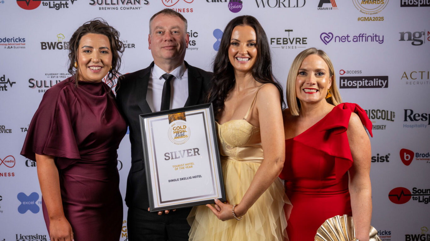 Four people dressed formally pose together, one holding a framed Silver award certificate, in front of a sponsor-branded backdrop at an awards event.