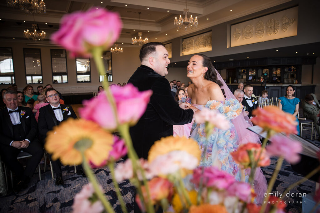 A couple stands smiling and holding hands during a wedding ceremony indoors, with colorful flowers in the foreground and guests seated in the background.