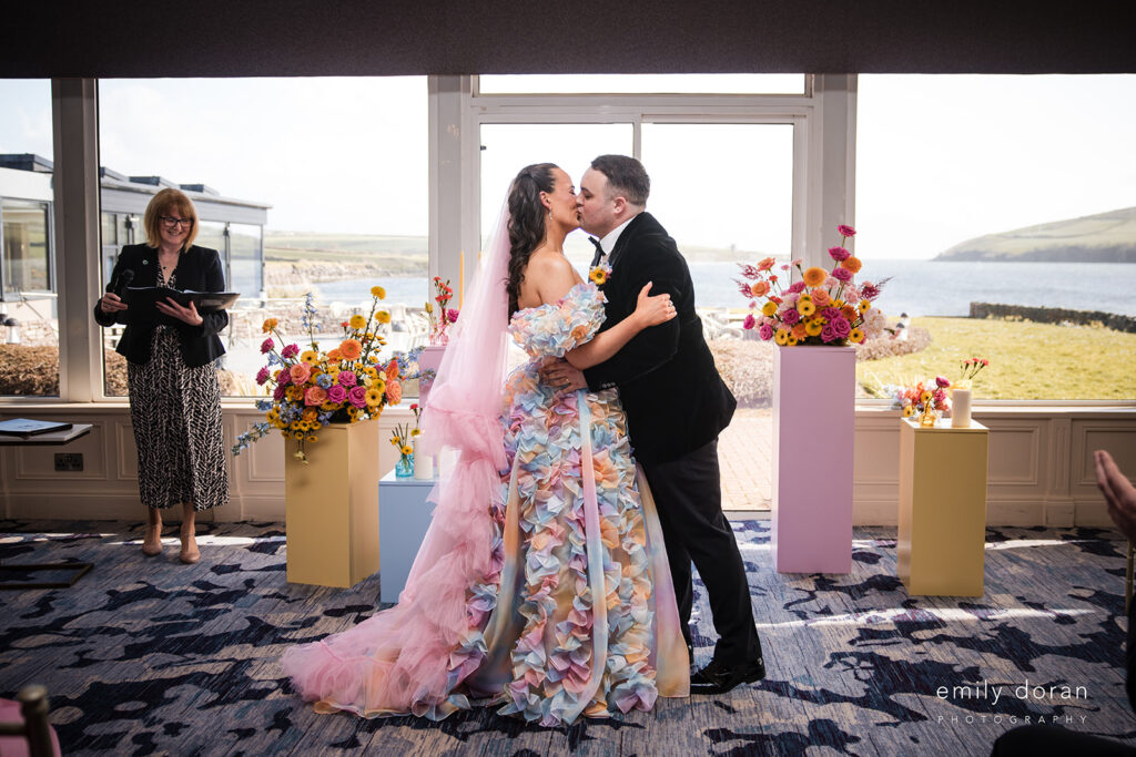 A bride in a colorful floral dress and a groom in a dark suit kiss at an indoor wedding ceremony, with floral arrangements and a scenic outdoor view in the background.