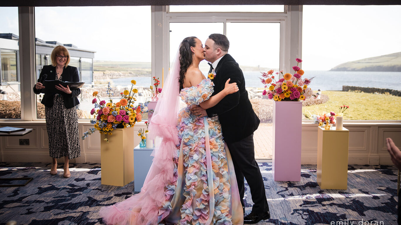 A bride in a colorful floral dress and a groom in a dark suit kiss at an indoor wedding ceremony, with floral arrangements and a scenic outdoor view in the background.