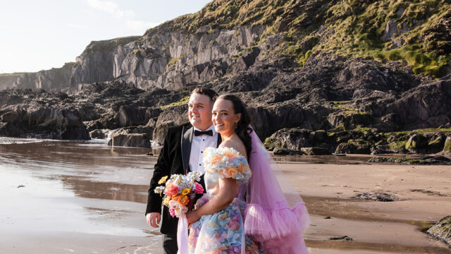 A couple stands on a beach in formal attire; the woman wears a colorful floral gown with a pink tulle veil, while the man wears a black suit and bow tie. Rocky cliffs rise in the background.