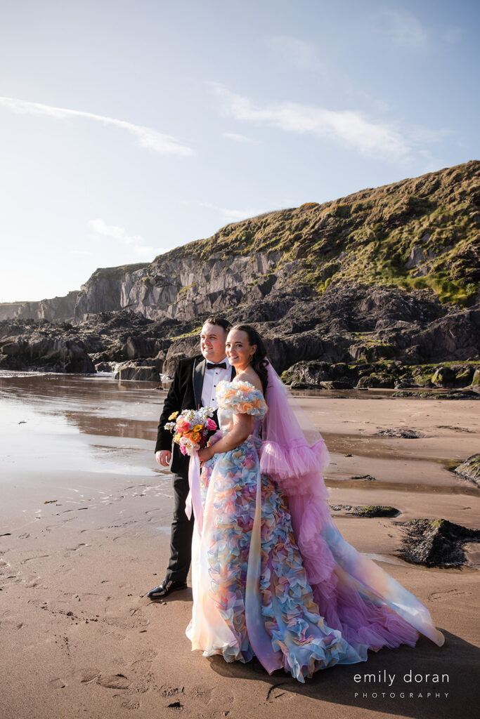A couple stands on a beach in formal attire; the woman wears a colorful floral gown with a pink tulle veil, while the man wears a black suit and bow tie. Rocky cliffs rise in the background.