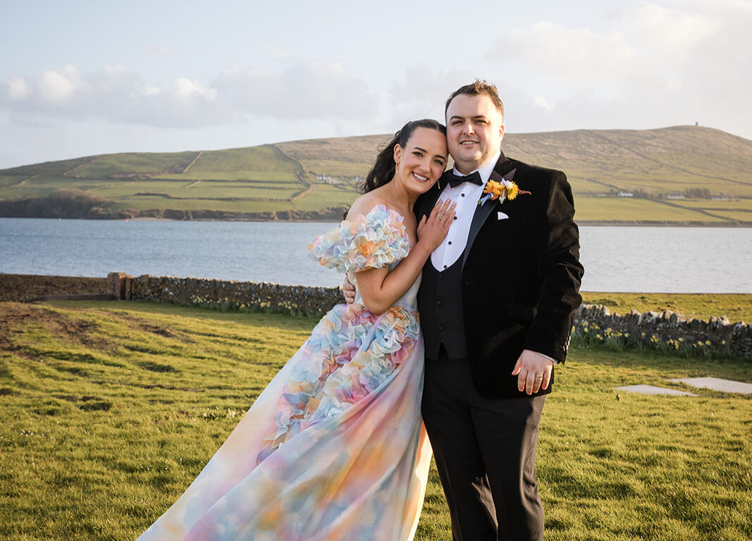 A couple dressed in formal attire stand smiling on grass near the ocean, with hills and blue sky in the background.