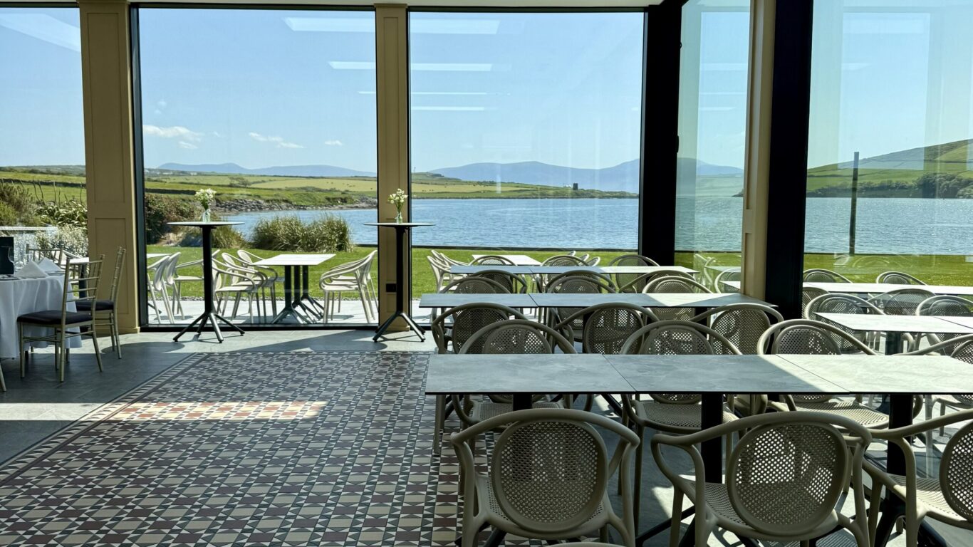 A glass-walled dining area with empty tables and chairs overlooks a scenic lake and green hills under a clear sky.